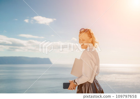 Freelance women sea working on a computer. Pretty middle aged woman with computer and phone outdoors with beautiful sea view. The concept of remote work. 109797658