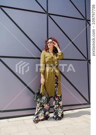 Portrait of a woman standing near a supermarket building. Caucasian model with long brunette hair, wears sunglasses and a khaki dress. Portrait of a woman standing near a supermarket building. Caucasian model with long brunette hair, wears sunglasses and a khaki dress. 109797786