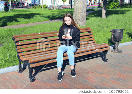 Young girl thoughtfully looking at smartphone sitting on bench in city park Young girl thoughtfully looking at smartphone sitting on bench in city park 109798661
