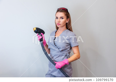 Professional cosmetologist woman holds gun from laser hair removal machine on gray background. Concept of painless hair removal Professional cosmetologist woman holds gun from laser hair removal machine on gray background. Concept of painless hair removal 109799170
