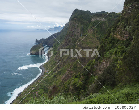Scenic elevated view towards the Rocha do Navio Reserve and Ponta de Catarina Pires cape. Ocean, tropical green vegetation, cliffs and waterfall. Portugal, Madeira, Santana. 109800027