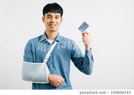Confident man smiles through a broken arm, using a credit card to cover emergency medical costs. Happy Asian man with sling support hand, isolated on white background, signifying quality health care. 109800243