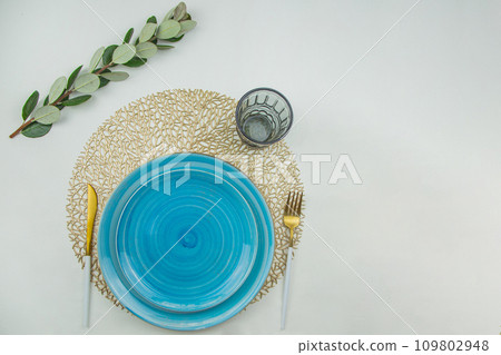 Two bright blue serving plates with a spiral pattern of different sizes, stand one on one on a golden textured napkin, gold - white serving devices are located on the side, a glass stands on top of Two bright blue serving plates with a spiral pattern of different sizes, stand one on one on a golden textured napkin, gold - white serving devices are located on the side, a glass stands on top of 109802948