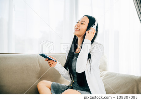 Attractive woman on sofa listens to music uses smartphone with headphones. Engaged in leisure relaxation and entertainment at home. Modern technology concept. Attractive woman on sofa listens to music uses smartphone with headphones. Engaged in leisure relaxation and entertainment at home. Modern technology concept. 109807093