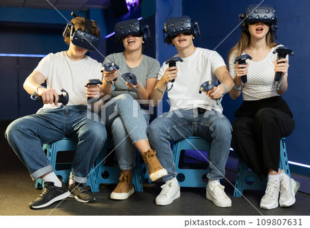 Girls and mans on virtual reality attraction sitting with another people and control joysticks Girls and mans on virtual reality attraction sitting with another people and control joysticks 109807631