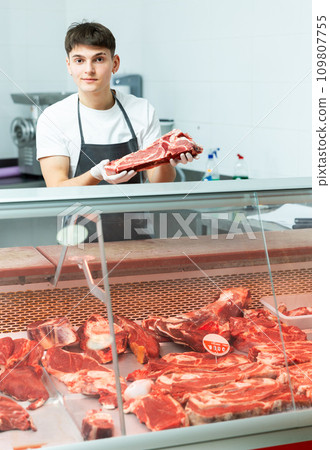 Young guy stands behind counter in meat department of supermarket and and holds piece of raw meat in his hands Young guy stands behind counter in meat department of supermarket and and holds piece of raw meat in his hands 109807755