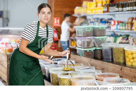Smiling salesgirl inviting to buy marinated olives in grocery store Smiling salesgirl inviting to buy marinated olives in grocery store 109807795
