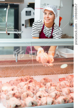 Young girl stands behind counter in meat department of supermarket and shows chicken carcass Young girl stands behind counter in meat department of supermarket and shows chicken carcass 109808057