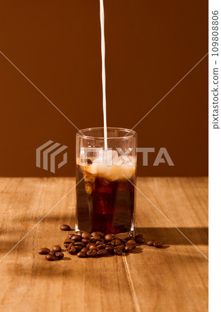 A stream of milk is pouring from above into a cup of black coffee on a wooden table with a brown background. Art space for food and beverage advertising. A stream of milk is pouring from above into a cup of black coffee on a wooden table with a brown background. Art space for food and beverage advertising. 109808806