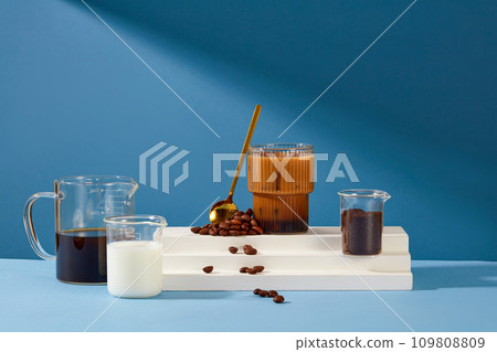 A glass of milky coffee is placed on a white platform with a metal spoon and coffee beans. Black coffee, fresh milk and coffee powder are stored in a beaker. Blue background for advertising. 109808809