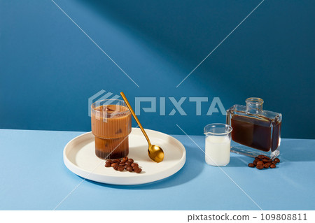 A cup of milk coffee is placed on a ceramic plate with a metal spoon and coffee beans, a cup of milk and a bottle of black coffee standing out against the blue background. 109808811