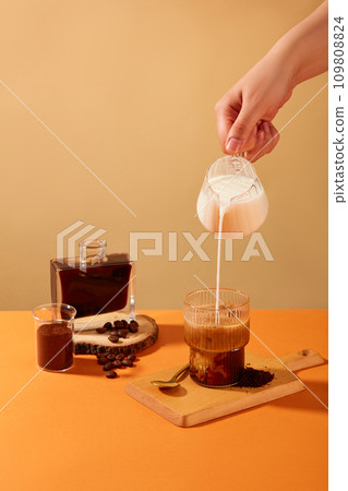 The hand is holding a glass of milk pouring into a cup of coffee, surrounded by coffee beans, coffee powder and black coffee. Coffee helps reduce the risk of Alzheimer's disease and dementia. The hand is holding a glass of milk pouring into a cup of coffee, surrounded by coffee beans, coffee powder and black coffee. Coffee helps reduce the risk of Alzheimer's disease and dementia. 109808824