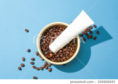 Close-up of a cosmetic tube placed on top of coffee beans in a ceramic bowl. Blue background for advertising. Coffee has antibacterial properties that help limit acne on the skin. Close-up of a cosmetic tube placed on top of coffee beans in a ceramic bowl. Blue background for advertising. Coffee has antibacterial properties that help limit acne on the skin. 109808843