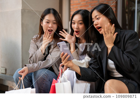 Excited female friends are looking at a phone screen with surprising faces, shocked with discount. 109809871