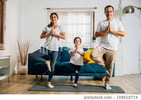 A family weekend at home becomes a harmonious and joyful experience as Asian parents and their daughter practice yoga and fitness promoting health happiness and togetherness. A family weekend at home becomes a harmonious and joyful experience as Asian parents and their daughter practice yoga and fitness promoting health happiness and togetherness. 109818719