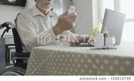 Senior man in wheelchair reading information about medication online, holding bottle of pills 109818799