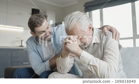 Son supporting upset senior father, family sitting on sofa at home, care Son supporting upset senior father, family sitting on sofa at home, care 109818849