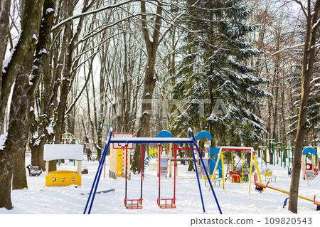 A quiet winter playground covered in snow, with colorful equipment and surrounded by bare evergreen trees. 109820543
