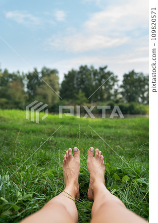The image shows a pair of bare feet lying on a grassy field with a lake and a green park in the background. Toes point upward. The image is peaceful and calm. The image shows a pair of bare feet lying on a grassy field with a lake and a green park in the background. Toes point upward. The image is peaceful and calm. 109820571