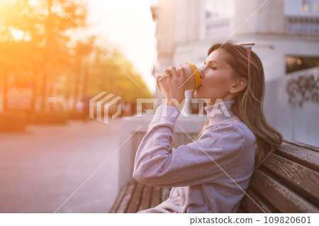 Woman drinks from cup on wooden bench. She is wearing a white shirt enjoying her beverage. The bench is located in a park setting, with trees in the background. 109820601