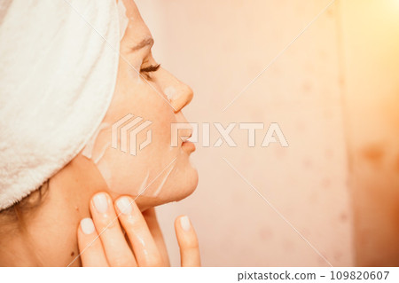 Young beautiful woman using a moisturizing facial mask after taking a bath. Pretty attractive girl in a towel on her head stands in front of a mirror in a home bathroom. Daily hygiene and skin care 109820607