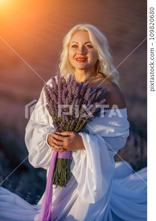 Blonde woman poses in lavender field at sunset. Happy woman in white dress holds lavender bouquet. Aromatherapy concept, lavender oil, photo session in lavender Blonde woman poses in lavender field at sunset. Happy woman in white dress holds lavender bouquet. Aromatherapy concept, lavender oil, photo session in lavender 109820680