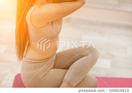 Young woman doing yoga in the gym. A girl with long hair and in a beige tracksuit stands in a cow pose on a pink carpet. A woman performs Gomukhasana. 109820819