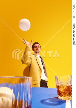 Young man, businessman in stylish suit paying ping pong, aiming ball into whiskey glass against bright yellow background 109821580