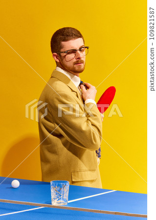 Handsome young man in stylish clothes playing tablet tennis, looking on empty whiskey glass against bright yellow background. Winner 109821587