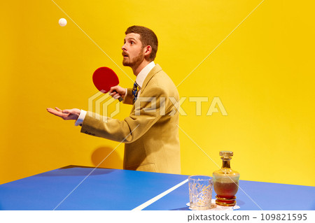 Handsome young man in elegant suit player table tennis against bright yellow background. Whiskey glass on table 109821595