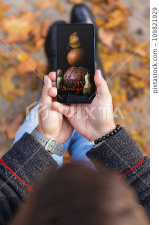 Man holding smartphone with Happy Thanksgiving photo message on the screen outdoors in a park full of autumn leaves on the ground 109821929