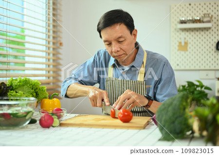 Middle age Asian man slicing tomatoes over a table for a vegan meal. Healthy lifestyle concept. Middle age Asian man slicing tomatoes over a table for a vegan meal. Healthy lifestyle concept. 109823015