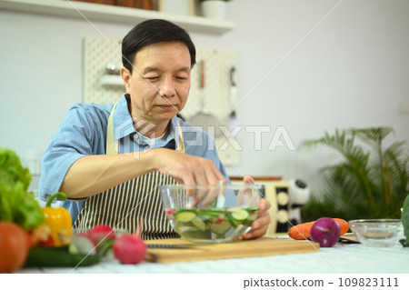 Smiling retired man making fresh vegetable salad, enjoying cooking alone at home. 109823111
