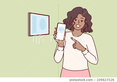 Woman holds mobile phone with thermometer standing near control panel for smart home 109823526