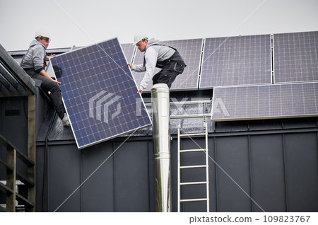 Men builders installing solar panel system on roof of house. Electricians in helmets lifting up photovoltaic solar module with help of ropes outdoors. Concept of alternative and renewable energy. Men builders installing solar panel system on roof of house. Electricians in helmets lifting up photovoltaic solar module with help of ropes outdoors. Concept of alternative and renewable energy. 109823767