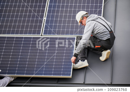 Man installer mounting photovoltaic solar panels on roof of house. Engineer in helmet installing solar module system with help of hex key. Concept of alternative, renewable energy. Man installer mounting photovoltaic solar panels on roof of house. Engineer in helmet installing solar module system with help of hex key. Concept of alternative, renewable energy. 109823768