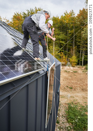 Men technicians installing solar panel system on roof of house. Workers in helmets lifting up photovoltaic solar module with help of ropes outdoors. Concept of alternative and renewable energy. Men technicians installing solar panel system on roof of house. Workers in helmets lifting up photovoltaic solar module with help of ropes outdoors. Concept of alternative and renewable energy. 109823771