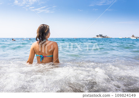 Attractive woman in bikini sitting on the beach and being splashed by the waves Attractive woman in bikini sitting on the beach and being splashed by the waves 109824149