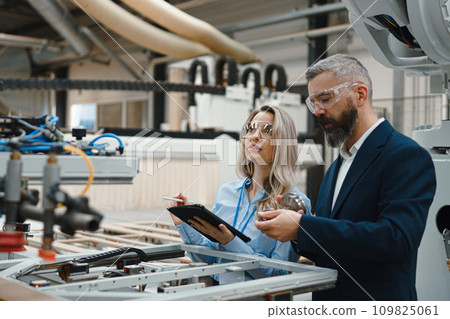 Female engineer and male project manager standing in modern industrial factory by precision robotic arm. Robot assembling wooden furniture in big furniture manufacturing facility with industrial Female engineer and male project manager standing in modern industrial factory by precision robotic arm. Robot assembling wooden furniture in big furniture manufacturing facility with industrial 109825061