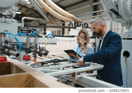 Female engineer and male project manager standing in modern industrial factory, talking about production of wooden furniture. Big furniture manufacturing facility with robotics and automation. Female engineer and male project manager standing in modern industrial factory, talking about production of wooden furniture. Big furniture manufacturing facility with robotics and automation. 109825063