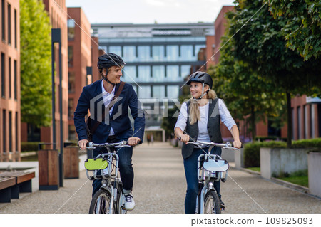 Spouses commuting through the city, riding bike on street. Middle-aged city commuters traveling from work by bike after a long workday. Spouses commuting through the city, riding bike on street. Middle-aged city commuters traveling from work by bike after a long workday. 109825093