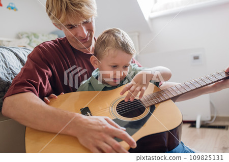 Father teaching boy to play on guitar. Son having fun in their room with dad, playing guitar and singing together. Concept of Father's Day, and fatherly love. Father teaching boy to play on guitar. Son having fun in their room with dad, playing guitar and singing together. Concept of Father's Day, and fatherly love. 109825131