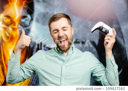 Young bearded man standing at the wall in the room and playing, holding wireless joystick, screaming, and winning intense video game match 109827162
