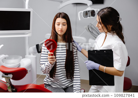 Young pretty girl looking sadly in the mirror while sitting in the dental chair, and the female doctor is encouraging the patient by holding her shoulder 109827841