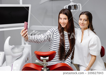 Young happy caucasian girl doing selfie shot on mobile phone, sitting at dental office chair with female dentist 109827843