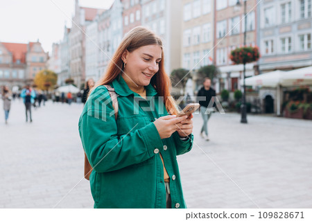 Happy cheerful young woman with backpack walking on city street checks her smartphone. Portrait of beautiful 30s girl using smartphone outdoors. Urban lifestyle concept. Happy cheerful young woman with backpack walking on city street checks her smartphone. Portrait of beautiful 30s girl using smartphone outdoors. Urban lifestyle concept. 109828671