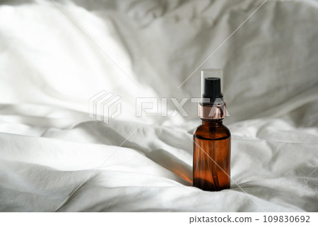 Cosmetic amber glass bottle with essential oil on white bed background with sunlights Cosmetic amber glass bottle with essential oil on white bed background with sunlights 109830692