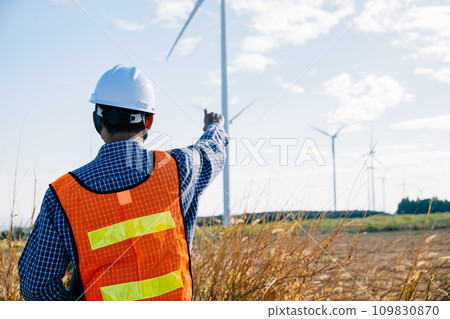 Engineer in hardhat checks windmill farm for clean energy. An Asian technician ensures turbine efficiency. Innovating for global electric security a portrait of expertise and dedication. 109830870