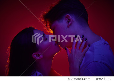 Portrait of young man and woman, loving couple kissing against red background in neon light. Anniversary Portrait of young man and woman, loving couple kissing against red background in neon light. Anniversary 109831289