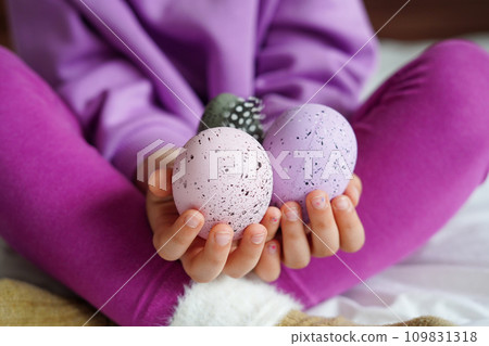 Hands of cute little child holding Easter eggs, child girl in purple and pink cloth sitting on the bed 109831318